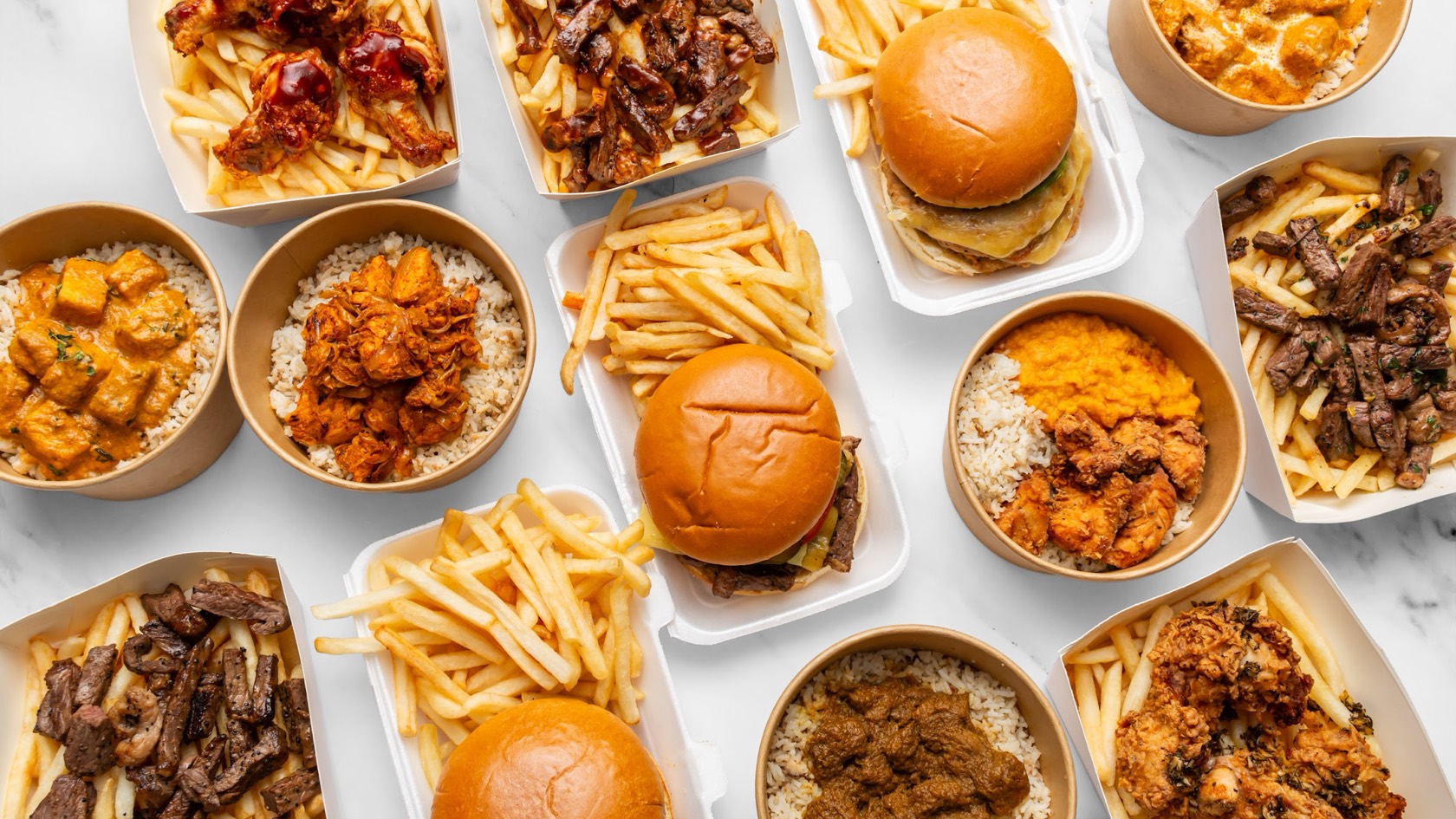 A full spread of Prime & Crisp burgers, steak boxes, wings, fried chicken and rice bowls laid out on marble